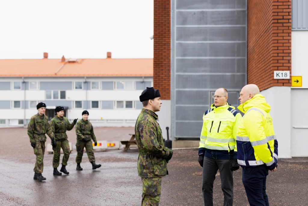 Kaksi keltatakkista Puolustuskiinteistöjen henkilöä keskustelee Puolustusvoimien sotilaan kanssa. Taustalla varusmiehiä ja kasarmi.